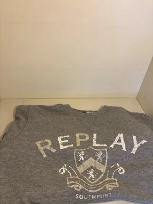 Grå sweatshirt från Replay - Snygg grå sweatshirt från Replay med tryckt logga och vapensköld på bröstet samt texten 'SOUTHPORT'. Tröjan har rund halsringning och är tillverkad i mjukt bomullsmaterial. Perfekt för en avslappnad och stilren look.