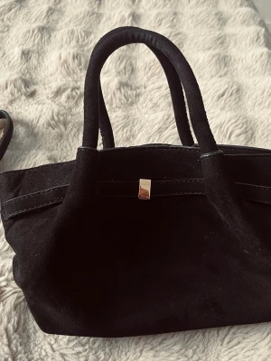 Svart handväska i mocka - Svart handväska – helt ny i perfekt skick. Väskan är rymlig och har gott om plats för allt som behövs i vardagen (rymmer en vattenflaska och mycket mer) och ger en lyxig känsla till din outfit. . Den har en avtagbar rem så att du kan bära den både i handen och över axeln