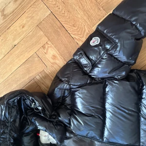 Svart Moncler pufferjacka med huva - Säljer en svart pufferjacka från Moncler med glansig finish och huva. Jackan har en ficka med Moncler-logga på ärmen, dragkedja framtill och quiltad design. Insidan har en cool serietidningsetikett. Perfekt för dig som vill ha en varm och snygg jacka med street vibe. Har att litet hål på under sidan av jackan som knappt är synligt.