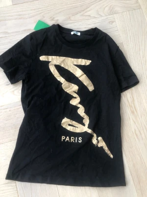 Svart Kenzo t-shirt med guldtryck - Kenzo t-shirt i svart med stort guldigt tryck och texten 'Paris' framtill. Klassisk rund hals och korta ärmar. Snygg och trendig design som sticker ut, perfekt för dig som gillar streetwear och exklusiva märken. Tillverkad i mjuk bomull för skön känsla.