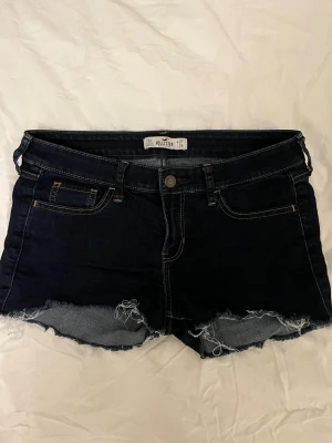 Mörkblå jeansshorts från Hollister - Snygga mörkblå jeansshorts från Hollister med råa, fransiga kanter och klassiska fem fickor. Shortsen har kontrastsömmar och en liten läderpatch med logga bak. Perfekta för sommaren och ger en avslappnad vibe.