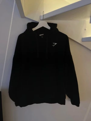 Svart hoodie från Gymshark - Snygg svart hoodie från Gymshark med deras logga tryckt på bröstet. Klassisk modell med huva och dragsnören samt ribbade muddar vid ärmar och nederkant. Perfekt för en avslappnad och sportig stil.