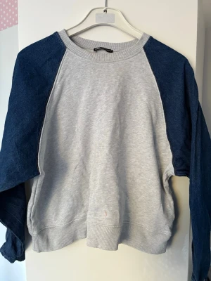 Grå sweatshirt med blå ärmar - Snygg sweatshirt med ljusgrå kropp och mörkblå raglanärmar. Tröjan har rund halsringning och ribbade muddar vid ärmslut och nederkant. Perfekt för en avslappnad och trendig look. Materialet känns mjukt och bekvämt.