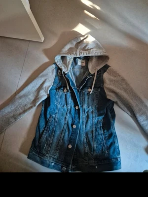 Jeansjacka med grå hoodie - Snygg jeansjacka från Jack & Jones. Jackan har vita knappar, två bröstfickor med lock och sidofickor. Perfekt för en avslappnad streetstil och enkel att matcha med olika outfits.