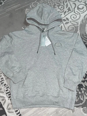 Grå hoodie från Dior med logga - Snygg grå hoodie från Dior med broderad CD-logga på bröstet. Klassisk modell med huva, dragsnören och magficka. Tillverkad i mjukt bomullsmaterial och har ribbade muddar vid ärmslut och nederkant. Perfekt för en chill och trendig look.