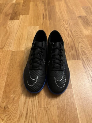 Nike mercurials svarta inomhus fotbollsskor med blå sula - Snygga fotbollsskor från Nike i svart med vita detaljer och stor vit Swoosh-logga. Skorna har blå yttersula och snörning för bra passform. Tillverkade i syntetmaterial med låg profil, perfekta för snabba rörelser på planen.
