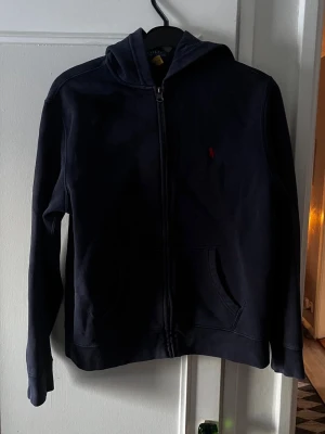 Mörkblå hoodie från Polo Ralph Lauren - Snygg mörkblå hoodie med dragkedja från Polo Ralph Lauren. Klassisk design med huva, två fickor framtill och den ikoniska röda loggan broderad på bröstet. Tillverkad i mjukt bomullsmaterial som är perfekt för en avslappnad stil. Storlek L(14-16). Säljer då den är för liten. Köpt här på plick.