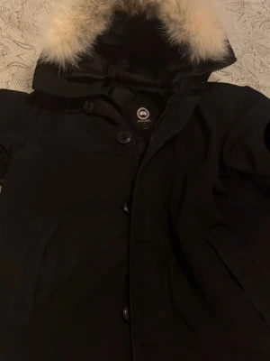 Svart parkas från Canada Goose XL - Säljer en svart parkas från Canada Goose med klassisk patch på ärmen och stor huva med fluffig, ljus päls. Jackan har dragkedja och knappar framtill samt flera fickor. Perfekt för kalla dagar och riktigt snygg streetstil. Mycket bra skick, inga skador.
