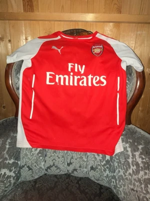 Arsenal matchtröja Puma röd/vit - Säljer en klassisk Arsenal matchtröja från Puma i rött och vitt med klubbmärke och Fly Emirates-tryck på bröstet. Tröjan har vita ärmar, rund hals och tryck med namnet BILLERSTAM samt nummer 15 på ryggen. Perfekt för dig som älskar fotboll och Arsenal.