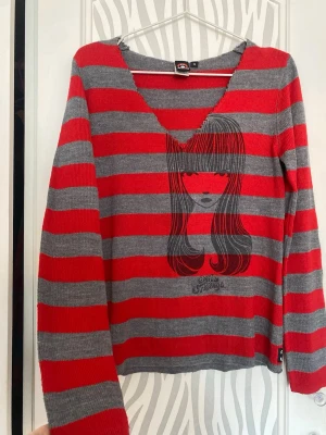 Röd och grå randig stickad tröja - Stickad tröja med breda röda och grå ränder från Emily the Strange. Tröjan har ett tryck av en tjej med långt hår på framsidan och en djup V-ringning. Långa ärmar och mjukt material. 
