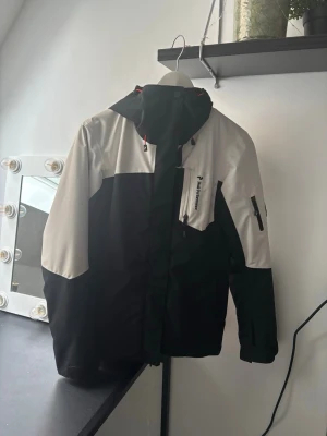 Svart och vit vindjacka från Peak Performance - Snygg svart och vit vindjacka från Peak Performance med huva och dragkedja. Jackan har fickor med dragkedja på bröstet och ärmen samt justerbar huva med röda detaljer. Perfekt för blåsiga dagar och outdoor-aktiviteter.