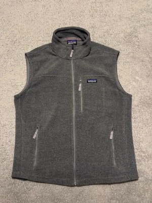Grå fleeceväst från Patagonia - Mörkgrå fleeceväst från Patagonia med hög krage och hel dragkedja framtill. Västen har tre fickor med dragkedja, en på bröstet och två på sidorna. Perfekt för lager-på-lager och enkel att matcha med olika outfits.