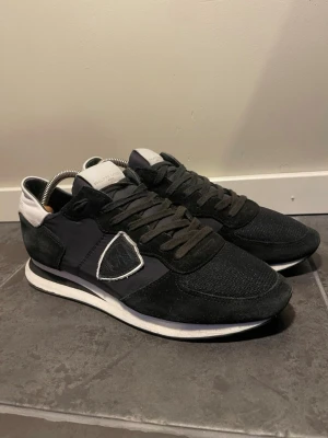 Svarta sneakers från Philippe Model - Snygga svarta sneakers från Philippe Model med vita detaljer och logga på sidan. Ovandelen är i en mix av mocka och textil, med svarta skosnören och vit sula. Skorna har en låg profil och en modern, sportig look.