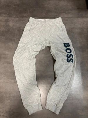 Grå mjukisbyxor från BOSS - Snygga grå mjukisbyxor från BOSS med elastisk midja och muddar vid bensluten. Byxorna har två sidofickor och en stor BOSS-logga i mörkblått längs ena benet. Perfekta för chill dagar eller när du vill ha en avslappnad stil.