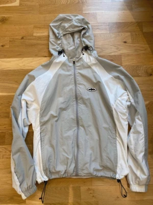 Corteiz vindjack/windbreaker  - Snygg vindjacka från Carhartt i ljusgrått och vitt med dragkedja framtill, huva med dragsko och elastiska muddar. Jackan har en sportig look och logga på bröstet. Perfekt för blåsiga dagar och enkel att matcha med olika outfits.
