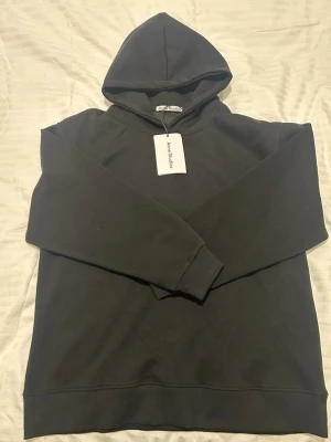 Svart hoodie från Acne Studios - Snygg svart hoodie från Acne Studios med tryck på ryggen där det står 'Acne Studios STOCKHOLM 1996'. Klassisk modell med huva och ribbade muddar. Perfekt för dig som gillar stilrena plagg med en touch av streetwear. Materialet känns mjukt och skönt.