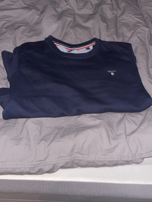 Mörkblå sweatshirt från GANT - Mörkblå sweatshirt från GANT med rund halsringning och diskret logga på bröstet. Tröjan har långa ärmar och ribbade muddar vid ärmslut och nederkant. Perfekt för en avslappnad och stilren look. Kan sänka pris vid snabb affär