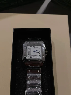  silverfärgad klocka - Stilren s klocka i silverfärgat rostfritt stål med fyrkantig boett och romerska siffror på vit urtavla. Länken har synliga skruvar och klockan har blå visare för en elegant touch. Automatisk rörelse och klassisk design.