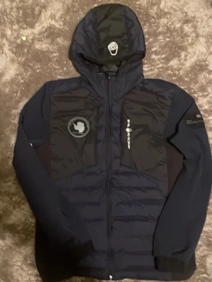 Svart Sail Racing vindjacka med huva - Snygg svart vindjacka från Sail Racing med huva och dragkedja. Jackan har Sail Antarctica Expedition-märke på bröstet och logga på ärmen. Materialet är lätt och vindtätt, perfekt för blåsiga dagar. Sportig design med paneler och diskreta detaljer.