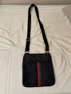 Svart axelväska från Gucci - Snygg svart axelväska från Gucci med justerbar axelrem. Väskan har det klassiska GG-mönstret och en röd-grön rand på framsidan. Materialet är canvas med läderdetaljer och dragkedja både fram och bak. Perfekt för dig som vill ha en stilren och ikonisk väska.