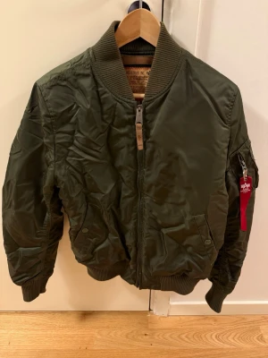 Olivgrön bomberjacka Alpha Industries - Snygg olivgrön bomberjacka från Alpha Industries med klassisk passform, ribbade muddar och dragkedja framtill. Jackan har fickor med lock och en ikonisk röd tag på ärmen. Tillverkad i slitstarkt nylonmaterial och har en cool militärinspirerad look.