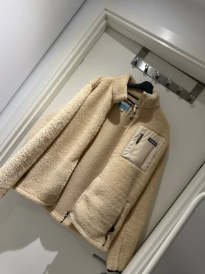 Beige teddyjacka från Columbia - Mysig beige teddyjacka från Columbia med hög krage och dragkedja. Jackan har en bröstficka med dragkedja och två sidofickor, alla med svarta detaljer. Tillverkad i fluffigt teddy-material som ger en avslappnad och trendig look.