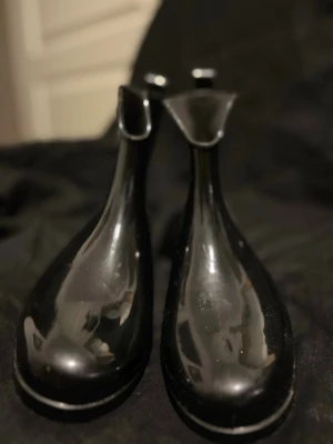 Svarta Chelsea boots i gummi - Snygga svarta Chelsea boots i glansigt gummi med robust sula och klassisk siluett. Perfekta för regniga dagar och har en stilren look som funkar till många outfits. Elastiska paneler på sidorna för enkel på- och avtagning.