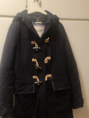 Marinblå duffelrock Hilfiger Denim XS - Klassisk marinblå duffelrock från Hilfiger Denim med huva och träknappar med repöglor. Rocken har två stora fickor framtill och är tillverkad i ett tjockare tyg som passar perfekt för kyligare dagar. Snygg och tidlös design med schyssta detaljer.