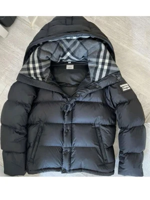Bubbery jacka - svart & grå/vit - storlek L - Snygg svart pufferjacka från Burberry med klassiskt rutigt foder i huvan och dragkedja framtill. Jackan har två sidofickor med dragkedja och tryckknappar, samt Burberry-logga på ärmen. Perfekt för kalla dagar.