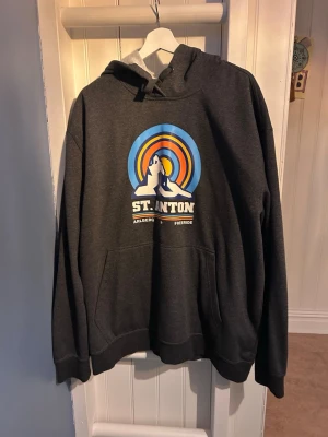 Mörkgrå St. Anton hoodie med tryck - Snygg mörkgrå hoodie från St. Anton med färgglatt tryck av berg och sol på bröstet. Texten 'St. Anton Arlberg Freeride' under motivet. Klassisk känguruficka framtill och justerbar huva med snören. Perfekt för dig som gillar streetwear och outdoor-vibe.