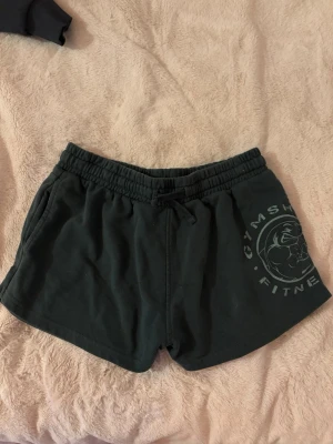  Gymshark shorts - Säljer ett par mörkgröna träningsshorts från Gymshark med elastisk midja och dragsko. Shortsen har fickor på sidorna och ett tryckt Gymshark Fitness-logotyp på vänster ben. Tillverkade i mjukt bomullsmaterial, perfekta för gymmet eller löpning. Använda ett fåtal gånger hemma men aldrig på gymmet. I fint skick. Orginalpris runt 400kr