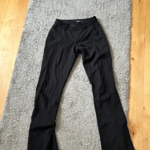 Svarta kostymbyxor med utsvängda ben - Snygga svarta kostymbyxor med utsvängda ben och enkel midja. Byxorna har en klassisk passform och är tillverkade i ett slätt tyg som ger en stilren look. Perfekta för dig som gillar en clean och tidlös stil.