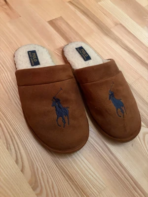 Polo Ralph Lauren tofflor - Bruna tofflor från Polo Ralph Lauren med blå broderad logga framtill. Utsidan är i mockaliknande material och insidan är fodrad med mjuk, vit teddy. Perfekta för att chilla hemma med stil. Ingen storlek står på tofflorna men sulan är ca 24,5-25cm.