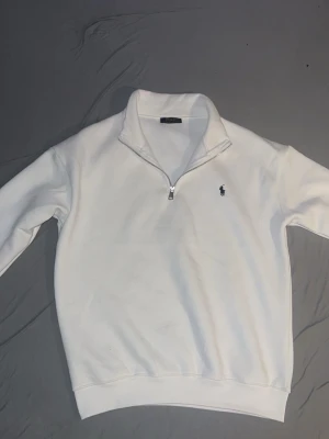 Vit quarter zip från Polo Ralph Lauren - Klassisk vit sweatshirt med quarter zip och hög krage från Polo Ralph Lauren. Tröjan har det ikoniska broderade logotypen på bröstet och ribbade muddar vid ärmslut och nederkant. Perfekt för en clean och stilren look.
