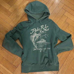 Grön hoodie Polo RL Key West - Grön hoodie från Polo Ralph Lauren med tryck på bröstet som visar en fisk, båt och texten 'Key West' och 'The Southernmost Point of U.S.'. Hoodien har dragsko i huvan och ribbade muddar.  Använd ett par gånger, inga fel på den