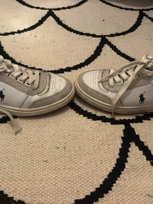 Vita sneakers från Polo Ralph Lauren - Snygga vita sneakers från Polo Ralph Lauren med klassisk låg siluett. Skorna har detaljer i grå mocka, vita skosnören och svart vadderad häl. Ikoniska Polo-loggan i mörkblått på sidan. Perfekta för en clean och sportig look.