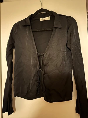 Svart blus från Pull&Bear - Trendig svart blus från Pull&Bear med lång ärm och öppen front som knyts med två snören. Perfekt för dig som gillar en stilren look. Blusen är skrynklig.
