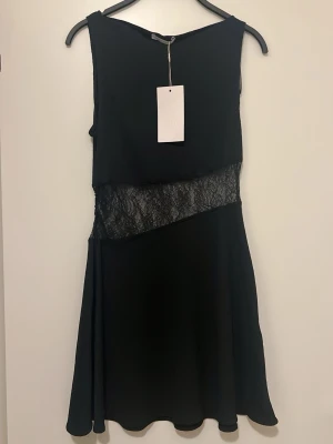 Svart klänning med spets från Zara - Snygg svart ärmlös klänning från Zara med en unik asymmetrisk spetsdetalj i midjan. Klänningen har en lätt utställd kjol och är gjord i ett tunt, mjukt tyg. Perfekt för dig som gillar stilrena plagg med en twist. Passar even till S