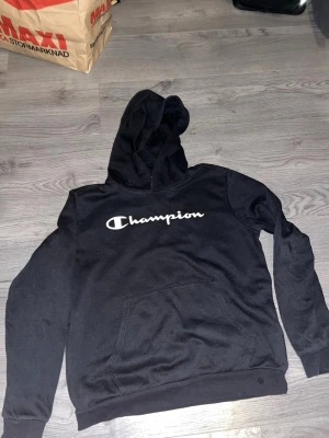 Svart Champion hoodie med logga - Svart hoodie från Champion med stor vit logga över bröstet. Klassisk känguruficka framtill och huva. Tillverkad i mjukt material som känns skönt mot huden. Perfekt för en avslappnad och sportig stil. Storlek 15-16 years old/ 174-179 cm som motsvarar S. Hör av dig vid frågor Mvh Edvin