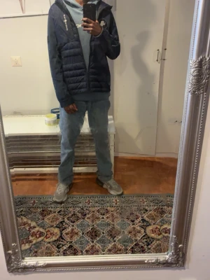 Mörkblå dunväst med huva - Mörkblå dunväst med huva och dragkedja framtill. Jackan har quiltad design och sidofickor, samt en liten logga på bröstet. Materialet är lätt och dunfyllt för extra värme. Perfekt för lager-på-lager och en clean streetwear-look.