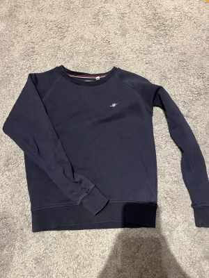 Mörkblå sweatshirt från GANT - Mörkblå sweatshirt från GANT med rund halsringning och liten broderad logga på bröstet. Tröjan har ribbade muddar vid ärmslut och nederkant. Tillverkad i mjuk bomullsblandning för en skön och avslappnad look.