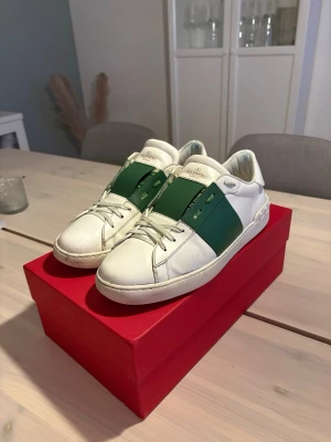 Valentino vita sneakers med grön detalj - Säljer ett par snygga vita och gröna sneakers från Valentino. Skorna är väldigt snygga och trendiga!, finns lite små defekter som att sulan är lite nedgången, lite sydda vid tungan men inte något mer, väldigt fint skick men använda. Tillverkade i läder för en exklusiv känsla och stilren look. Priset är inte hugget i sten, pris kan diskuteras vid snabb affär!