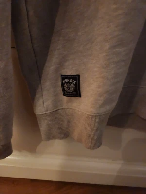 Beige hoodie från Morris - Säljer en beige hoodie från Morris med dragsko i huvan och ribbade muddar. Klassisk passform och liten Morris-logga på bröstet samt en patch vid nederkanten. Perfekt för dig som gillar stilrena och bekväma plagg.