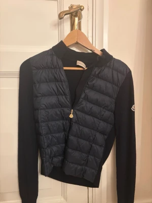 Mörkblå cardiganjacka från Moncler - Snygg mörkblå cardiganjacka från Moncler med quiltad front och stickade ärmar. Jackan har dragkedja framtill och en liten Moncler-logga på ärmen. Perfekt för våren och hösten. (Den är i barnstorlek men jag som är 167 passar den för men lite tajt runt magen💗) Skulle säga att den passar någon som har xs. Den är använd men de syns knappt förutom att moncler loggan vid nacken har lostnat lite! Har tyvärr inte kvar äktahetsbevis men skickar gärna när jag screenar qr koden igen😊