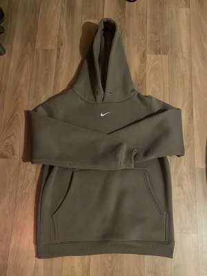   Nike hoodie - Snygg brun hoodie från Nike med klassisk vit swoosh-logga på bröstet. Hoodien har stor känguruficka framtill, ribbade muddar och en rejäl huva med dragsko. Perfekt för en chill och sportig look.