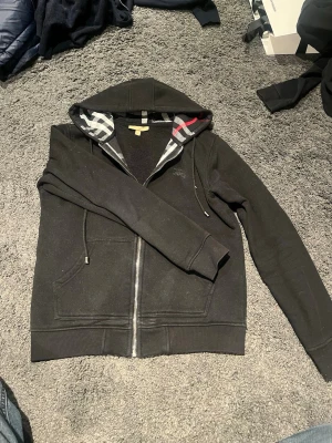 Svart Burberry hoodie med dragkedja - Svart hoodie från Burberry med dragkedja framtill och klassiskt rutigt foder i huvan. Två fickor fram och ribbade muddar vid ärmslut och nederkant. Tillverkad i mjukt bomullsmaterial och har justerbar dragsko i huvan. Perfekt för en avslappnad och stilren look.