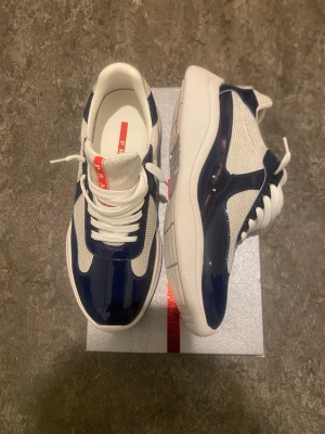Prada Cups - Snygga sneakers från Prada i en kombination av marinblått och vitt. Skorna har en chunky sula, vita skosnören och detaljer i glansigt material samt mesh-tyg på sidorna. Perfekta för dig som vill ha en stilren och modern look. Kommer med originalboxen
