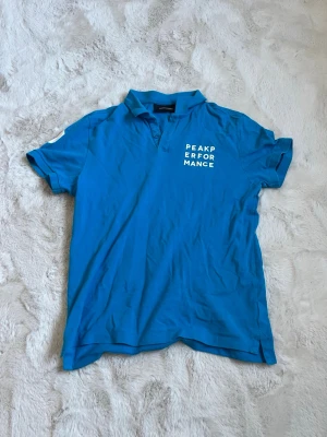 Blå Peak Performance t-shirt - Snygg blå t-shirt från Peak Performance med vit logga på bröstet. Klassisk passform med korta ärmar och krage. Perfekt för en avslappnad stil och enkel att matcha med jeans eller shorts.
