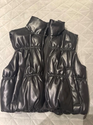 Svart pälspuffväst från NA-KD XS - Snygg svart pälspuffväst från NA-KD i storlek XS. Västen har glansig yta, hög krage och dragkedja framtill. Den är vadderad och har två fickor på sidorna. Perfekt för lager-på-lager och trendig streetstyle.