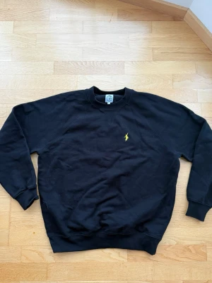 Svart sweatshirt från Polar Skate Co. - Klassisk svart ocersize sweatshirt från Polar Skate Co. med gul broderad logga på bröstet. Tröjan har rund halsringning, ribbade muddar vid ärmslut och nederkant samt en avslappnad passform. Perfekt för en casual look och häng i skateparken. 